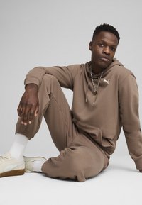 Sudadera marrón y pantalones de chándal a juego, hechos de una tela suave, con un bolsillo tipo canguro y acento de logo. Zapatillas blancas con suelas beiges.