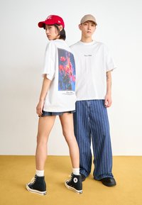 Hvite overdimensjonerte t-skjorter med et fargerikt blomsterdesign på baksiden, kombinert med mørke denimshorts og stripete vide bukser. Svarte sneakers.