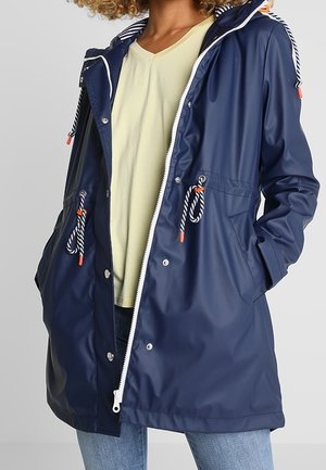 Personne portant une veste imperméable bleu marine à cordons rayés, un t-shirt jaune et un jean, debout les mains dans les poches.