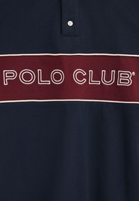 Granatowa koszulka polo z bordową tasiemką z napisem "POLO CLUB" w białym obrysie, krótkie rękawy, zapięcie na guziki pod kołnierzykiem oraz gładką bawełnianą teksturą.