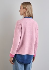Maglione rosa con una finitura testurizzata, dalla vestibilità ampia, con polsini rosa, indossato sopra una camicia di jeans con orlo arrotondato.