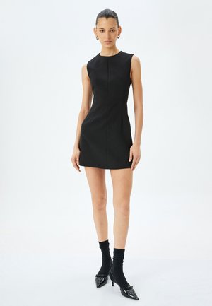 Robe noire sans manches avec une silhouette ajustée, longueur au-dessus du genou, texture lisse et lignes épurées. Associée à des chaussettes noires et des talons pointus.