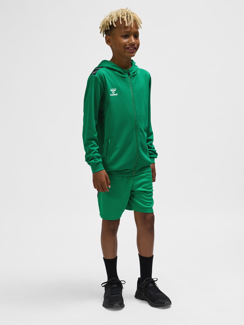 Chłopiec z blond dredami noszący zieloną bluzę z kapturem na zamek i pasujące szorty, stojący w czarnych skarpetkach i czarnych trampkach na jednolitym tle.