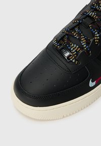 Bout de chaussure de sneaker en cuir noir avec perforations, lacets multicolores avec des éclaboussures, swoosh rouge et blanc brodé, et semelle blanc cassé sur fond blanc.