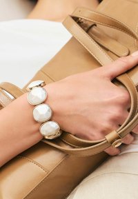 Beige læderhåndtaske holdt i en hånd, med et guld-accenteret armbånd med fire hvide, facetterede sten. Glat tekstur og stilrent design.