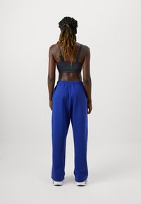 Melna sporta krūšturis un brīvi pieguļošas zilas treniņbikses ar elastīgu jostasvietu, redzami no aizmugures, uz personas ar pīnes.