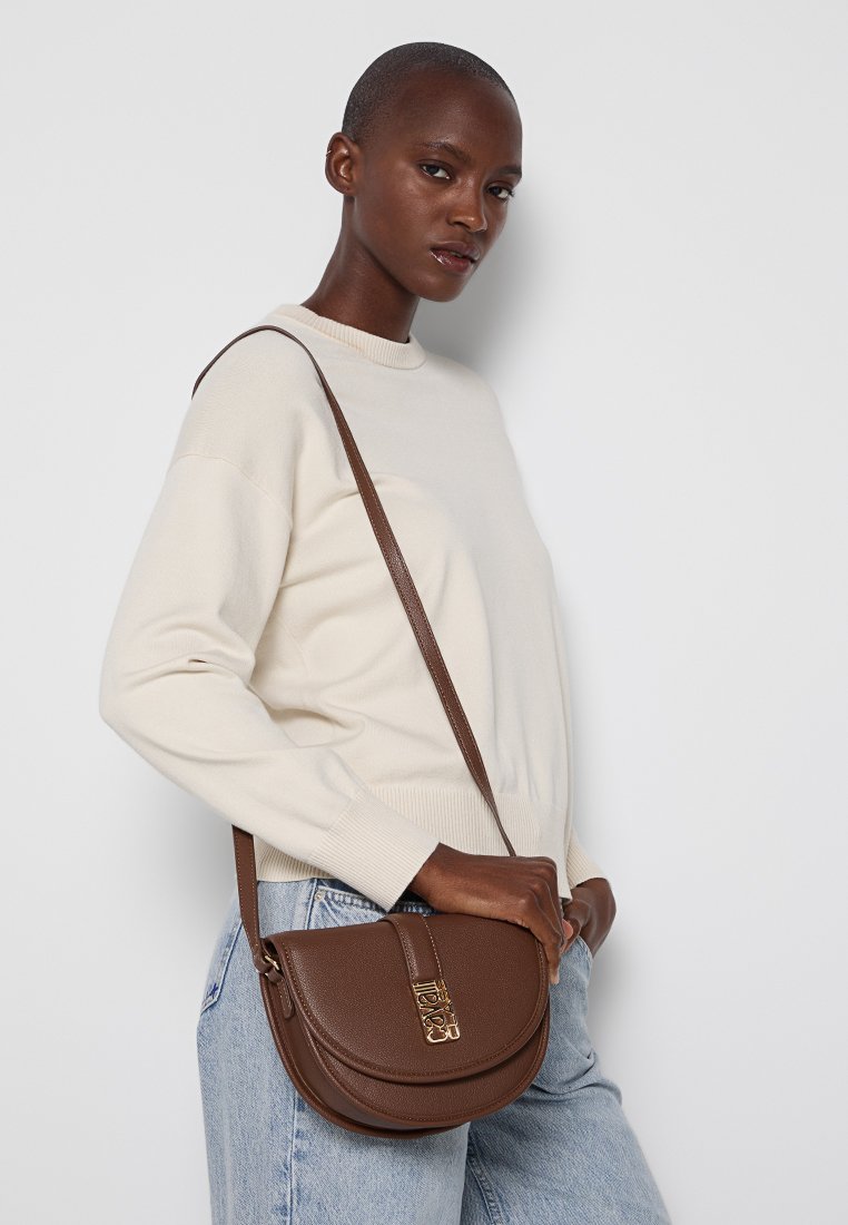 Bolso cruzado de cuero marrón con forma curvada, acentos de hardware dorado y un acabado texturizado, llevado sobre un suéter crema y jeans azules.