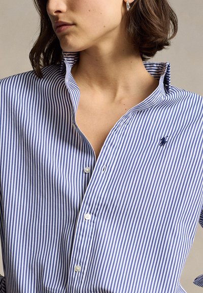 Mujer con una camisa azul y blanca de rayas verticales con botones, cuello levantado y pequeño logo bordado en el pecho.