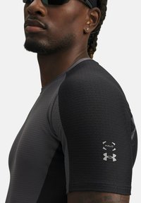 Mann in eng anliegendem schwarzen und grauen Sportshirt mit Under Armour und Halo-Logos auf dem Ärmel, Sonnenbrille und Ohrringen.