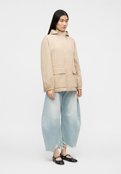 Γυναίκα που φοράει μπεζ μπουφάν με κουκούλα, ανοιχτό μπλε τζιν παντελόνι με φαρδιά πόδια και σκισίματα, και μαύρα παπούτσια Mary Jane με μεταλλικές λεπτομέρειες, στέκεται μπροστά σε λευκό φόντο.