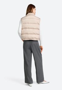 Beige puffer gilet met een hoge kraag, gestikt ontwerp en geribbelde trui-kraag. Gecombineerd met grijze wijde broek en witte platte schoenen.