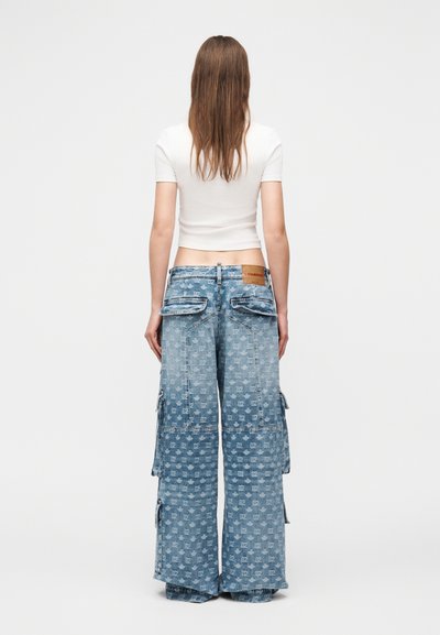 Žena s dugom smeđom kosom nosi bijelu kratku majicu s kratkim rukavima i široke svijetloplave cargo traperice s printom po cijelom kroju, snimljena s leđa.