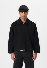 Svart zip-up jakke med spiss krage, frontlommer og brodert gult logo på venstre bryst. Har en avslappet passform og et robust stoff.