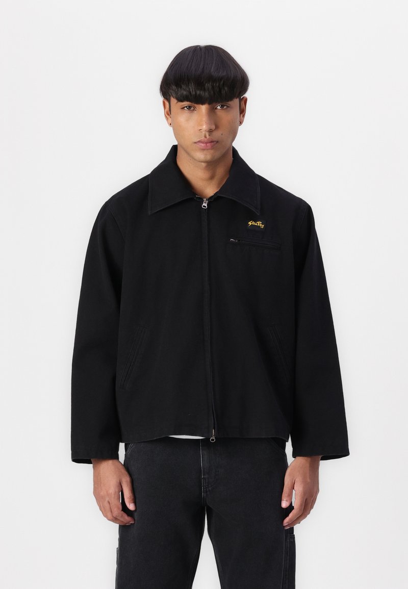 Juoda zip-up striukė su smailiu apykakle, priekinių kišenių ir siuvinėtu geltonu logotipu kairėje krūtinės pusėje. Savybės: laisvas kirpimas ir tvirta medžiaga.
