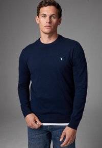 Maglione lavorato a maglia blu navy con maniche lunghe, scollo rotondo e polsini a coste. Presenta un piccolo emblema azzurro sul petto.