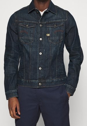 Homme portant une veste en denim bleu foncé avec boutons sur le devant et poches poitrine à rabat, associée à un pantalon bleu marine et un sous-vêtement blanc visible au col.