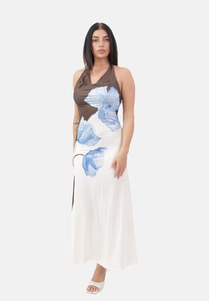 Mujer vestida con un vestido de tirantes marrón y blanco con grandes estampados de flores azules, de pie frente a un fondo claro y sencillo, llevando sandalias blancas.