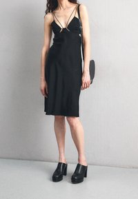 Černé šaty bez rukávů s tenkými ramínky, se střiženými detaily na hrudi. Spárované s robustními černými platformovými muly a malou taškou.
