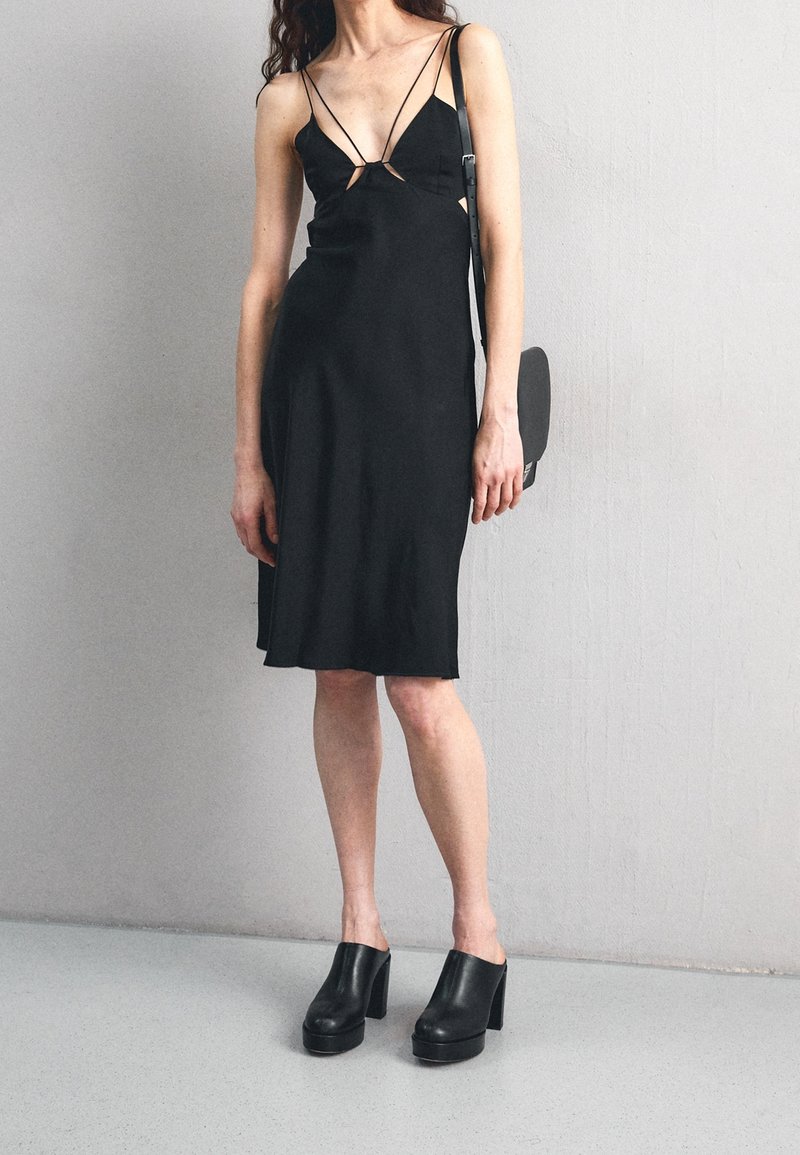 Černé šaty bez rukávů s tenkými ramínky, se střiženými detaily na hrudi. Spárované s robustními černými platformovými muly a malou taškou.