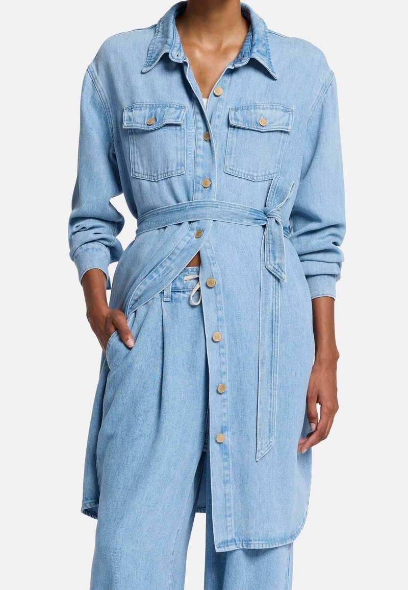 Femme portant une veste ceinturée en jean bleu clair avec boutons, poches poitrine à rabat, et un pantalon large assorti en jean.