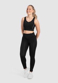 Schwarzer Sport-BH mit Reißverschluss und reflektierenden Akzenten, kombiniert mit hoch taillierten schwarzen Leggings. Gestylt mit weißen Sportschuhen.