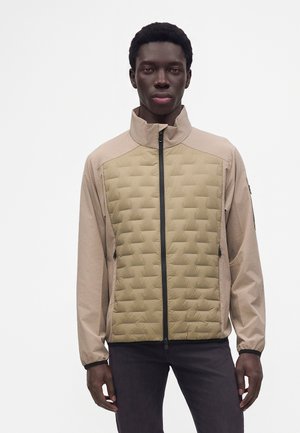 Mann trägt beige gesteppte Jacke mit Reißverschluss und steht vor einfachem weißem Hintergrund.