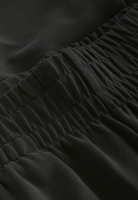 Černá tkanina s hladkým povrchem obsahuje nařasené elastické detaily, které vytvářejí zpevněný efekt podél okraje a předvádějí vlnitý design.