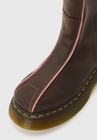 Pruun nahast saabas, mille õmbluste ääres on roosad triibud ja kollased õmblused, mis ühendavad läbipaistva kollase taldri.