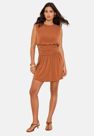 Młoda kobieta z długimi ciemnymi włosami ubrana w bezrękawnikową krótką sukienkę w kolorze rdzy, złote kolczyki koła, bransoletki i brązowe sandały na obcasie, stoi.