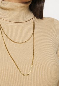 Colliers en chaîne couleur or superposés sur un pull à col roulé côtelé beige. La texture du pull est douce avec une coupe ajustée.