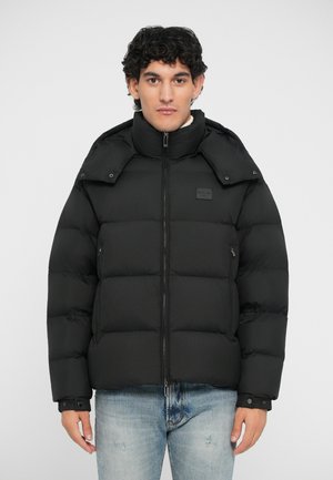 Schwarze Daunenjacke mit hohem Kragen, Reißverschluss und geknöpften Manschetten. Besitzt ein gesteppte Design und ein kleines Logo-Patch auf der Brust.