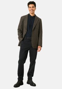 Brauner Hahnentrittmuster-Blazer über einem marineblauen Hemd mit Knopfleiste, kombiniert mit schwarzen Hosen und schwarzen Stiefeletten. Glatte Textur mit taillierter Passform.