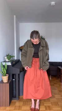 Woman adjusting rust-colored skirt, wearing olive green jacket and black top, standing in living room with plants and piano.
