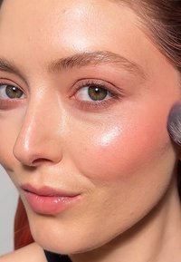 Close-up van een gezicht met een lichte huid, een gladde textuur, natuurlijke makeup, een zachte roze blush op de wangen en een glanzende lipfinish. Bruine ogen en donkere wenkbrauwen.