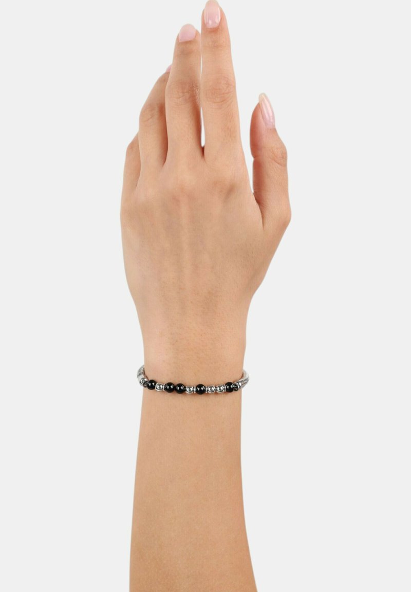 Mano con unghie naturali che indossa un braccialetto d'argento con perline alternate nere e argento su uno sfondo bianco semplice.
