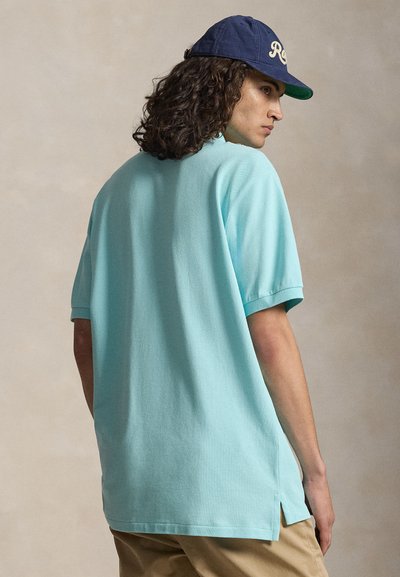 Junger Mann mit lockigem Haar, der eine blaue Baseballkappe, ein hellblaues Kurzarmhemd und beige Hose trägt, leicht zur Seite gedreht.