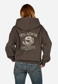 Barna kapucnis pulóver, amelyen egy k�ávéscsésze grafikai tervezése és a "PEGADOR COZY NIGHTS" felirat látható a hátsó részen. Az anyag lágynek tűnik, lazán illeszkedik.