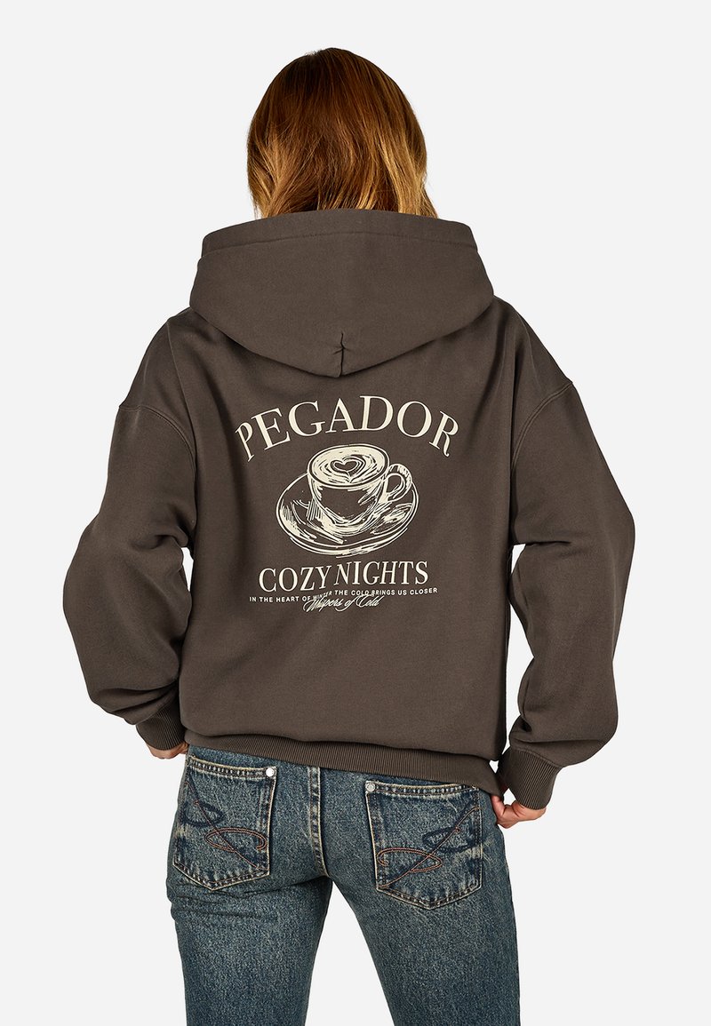Barna kapucnis pulóver, amelyen egy kávéscsésze grafikai tervezése és a "PEGADOR COZY NIGHTS" felirat látható a hátsó részen. Az anyag lágynek tűnik, lazán illeszkedik.