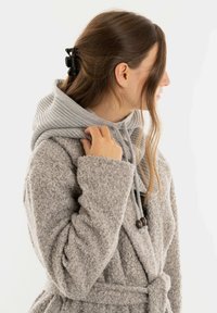 Vrouw in zijprofiel met een gestructureerde grijze jas over een gebreide hoodie, die de kraag van de hoodie vasthoudt met haar hand, haar haar opgestoken.