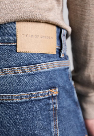 Jeans en denim présentant une couleur bleu clair, des coutures visibles en orange et une patch en cuir beige rectangulaire étiquetée "TIGER OF SWEDEN."