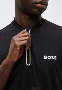 Άνδρας με σκουρόχρωμο δέρμα που κλείνει μια μαύρη μπλούζα BOSS με κάθετη ριγωτή λεπτομέρεια φερμουάρ κοντά στον γιακά.