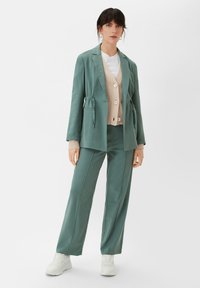 Helles grünes Blazer mit einer Krawatte in der Taille, kombiniert mit einem beigen, knöpfbaren Cardigan und passenden Hosen. Weiße Sneaker runden das Outfit ab.