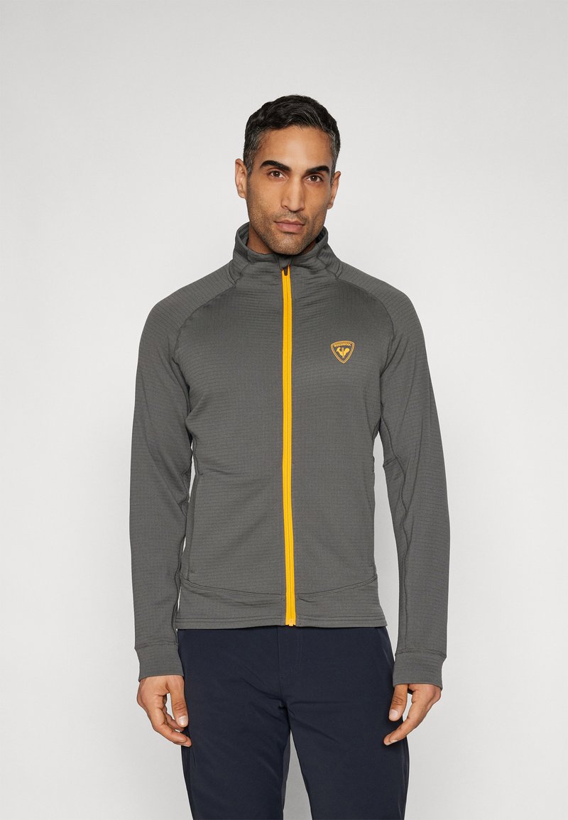 Mężczyzna w szarej kurtce zapinanej na zamek z żółtym suwakiem i logo, stojący na jednolitym jasnym tle.