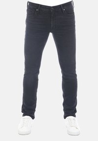 Jeans de mezclilla negros con un corte ajustado, diseño de cinco bolsillos y detalles de costura sutiles, combinados con zapatillas blancas sobre un fondo claro.