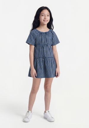 Ung pige med langt mørkt hår iført en blå og hvid ternet kortærmet kjole og hvide sneakers står foran en ensfarvet hvid baggrund.