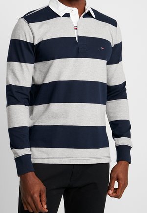 Torse masculin portant un maillot de rugby à larges rayures horizontales bleu marine et gris, avec un col blanc et une patte de boutonnage, assorti à un pantalon noir.