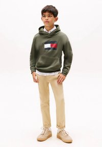 Groene hoodie met een centrale grafische afbeelding in marineblauw, rood en wit, gecombineerd met beige broek en lichtgekleurde sneakers.