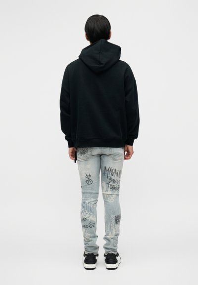 Μαύρο φούτερ με κουκούλα, άνετη εφαρμογή; ανοιχτό μπλε τζιν με σχέδια γκράφιτι; μαύρα και λευκά αθλητικά παπούτσια; θέα από πίσω.