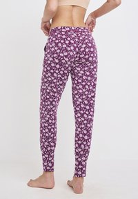 Kobieta w purpurowych spodniach w kwiatowy wzór i beżowym topie na krótki rękaw, pokazana z tyłu, stojąca boso na tle jednolitym.