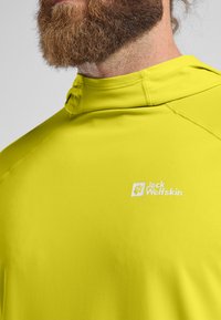 Maglietta sportiva verde lime a maniche lunghe con colletto alto. Presenta un logo bianco sul petto e un tessuto liscio traspirante che assorbe l'umidità.